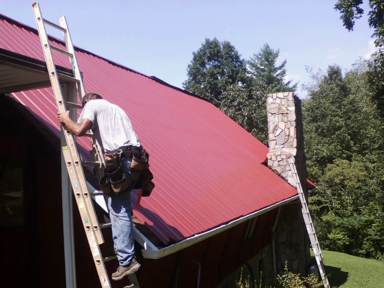 Roofer on a ladder beside a red metal roof and stone chimney — Soffit & Fascia Repair by Roofing Brothers in Fiskdale